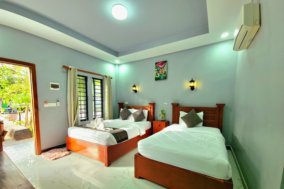 A hotel room with two beds, a large window with curtains, a wall-mounted air conditioner, and a small bedside table with a plant.