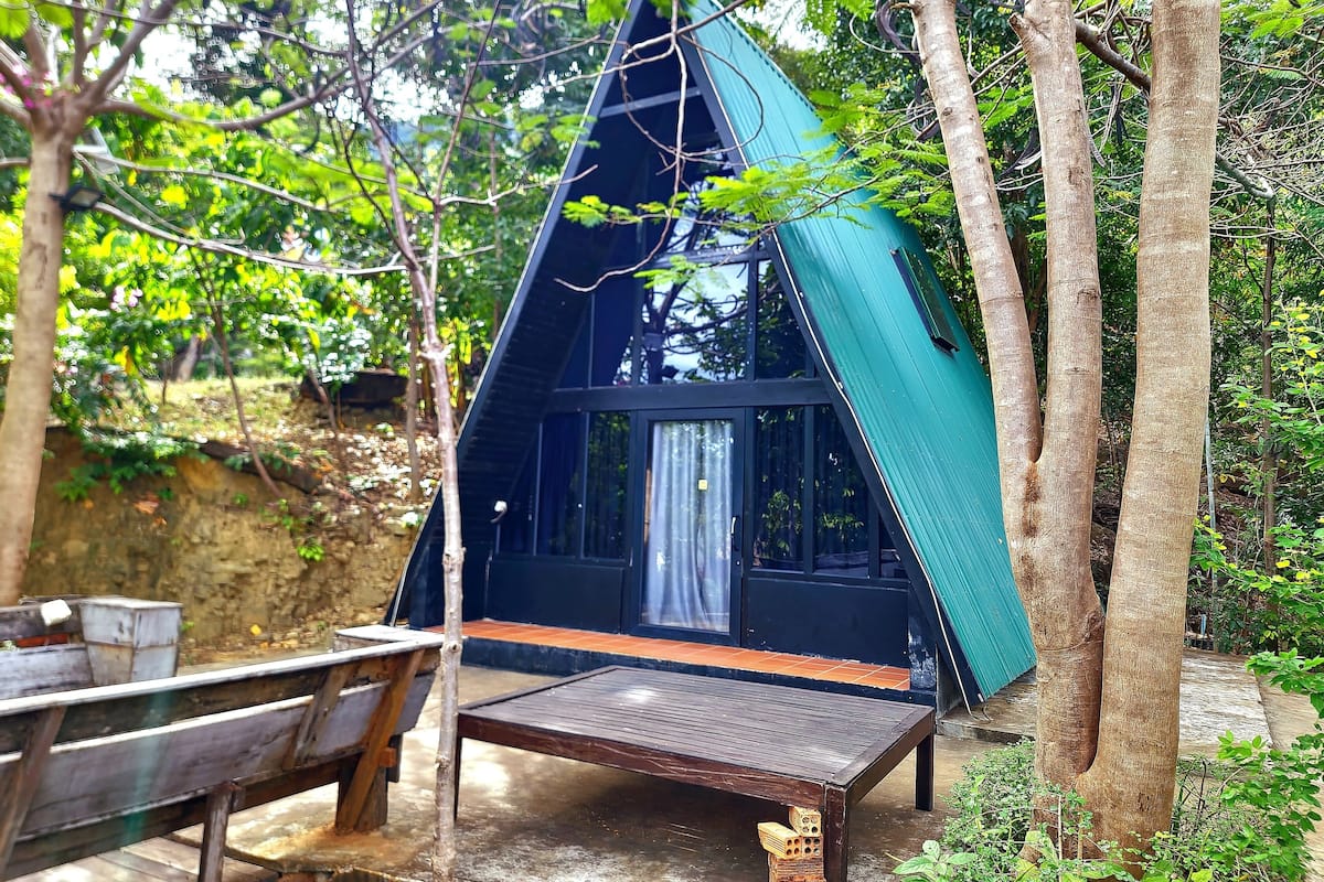 A small, dark-colored, triangular-shaped cabin with a wooden bench and a table in front, surrounded by trees.