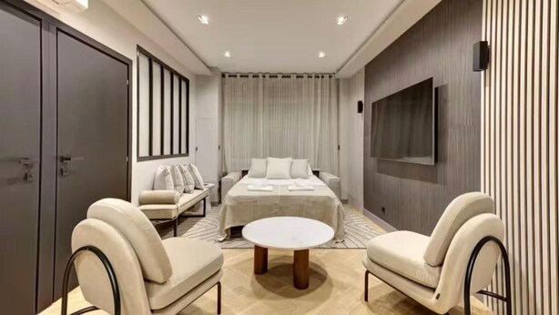 Una habitación de hotel moderna con una cama grande, un sofá, dos sillones, una mesa de centro redonda y un televisor montado en la pared.