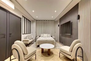 A modern hotel room with a large bed, a sofa, two armchairs, a round coffee table, and a TV mounted on the wall.
