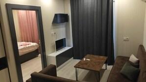 A compact living space with a sofa, a coffee table, a TV mounted on the wall, and a bed with curtains. - pozcu forum plus (Mersin)