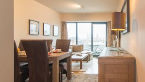 A modern hotel room with a dining area, a sofa, a coffee table, and a view of the cityscape through large windows. - Apartment, few Meters From Beach (Ostend)