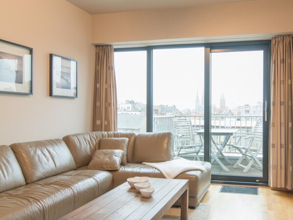 A modern living room with a leather sofa, a wooden coffee table, and a city view through large windows. - Apartment, few Meters From Beach (Ostend)