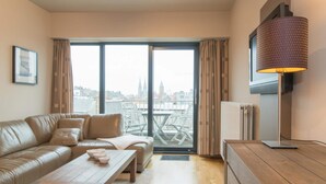 A modern living room with a leather sofa, wooden coffee table, and a view of a cityscape through large windows. - Apartment, few Meters From Beach (Ostend)