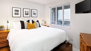 A bedroom with a large bed, a nightstand with a lamp, a dresser, and a TV mounted on the wall. There are three framed pictures on the wall above the bed.