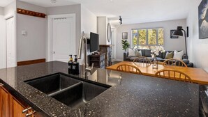 A modern kitchen with a black granite countertop and a double sink. There is a dining area with a wooden table and chairs, and a living room with a sofa and a large window.