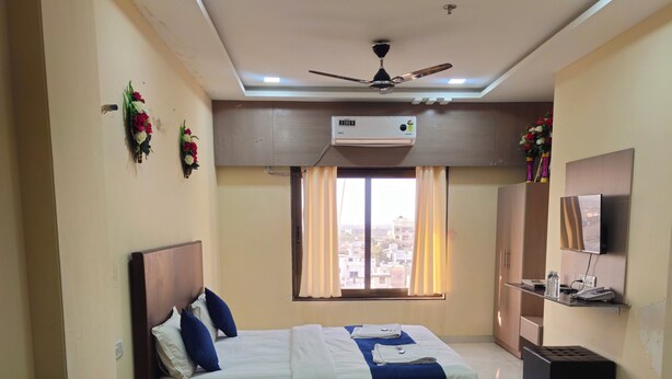 A hotel room with a bed, a ceiling fan, an air conditioning unit, a window with curtains, a TV, and a small table with a bottle and a phone. - Hotel Lord Krishna Dham (Dwarka)