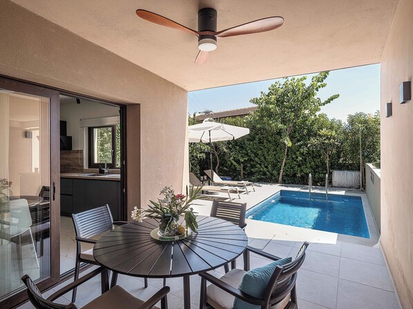 A modern outdoor dining area with a pool, surrounded by greenery and a clear sky.