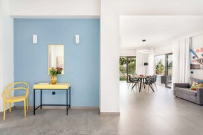 A modern living space with a blue accent wall, a yellow chair, a desk with a vase of flowers, and a dining area with a table and chairs. - Villa Fos | Private pool, elegance & seaview! (Crete)
