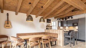 A modern kitchen with a wooden ceiling, a dining area with a wooden table and cushioned chairs, and a kitchen island with bar stools. - Superior Ferienhaus # 4A mit Sauna (Uttendorf)