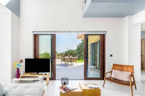 A modern living room with a flat-screen TV, a sofa, a coffee table, and a dining area with outdoor seating. - Villa Alya | Heated* pool, luxury & seaviews (Crete)