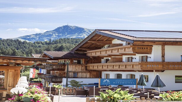 A traditional wooden building with a sign for 'Alps Kitzbühel' hotel, outdoor seating, and a mountain view.