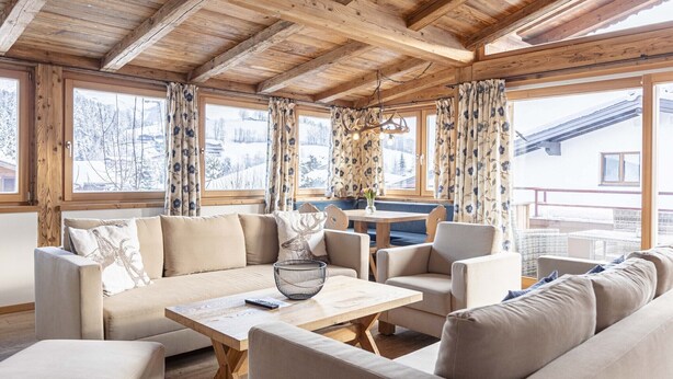 A cozy room with wooden beams, a sofa, chairs, a table, and large windows with curtains.