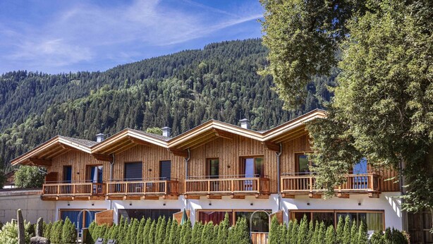 A wooden chalet with a balcony, surrounded by a well-maintained lawn and trees, set against a mountainous backdrop.