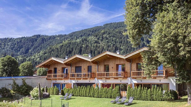 Un chalet in legno con diversi balconi, un giardino curato con lettini prendisole e uno sfondo montuoso.