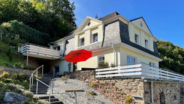 A two-story house with a stone retaining wall, a red umbrella, and a staircase leading to a balcony. - Vacation apartment Am Kreuzberg Apartment No. 3 (Nordrhein-Westfalen - Schleiden)