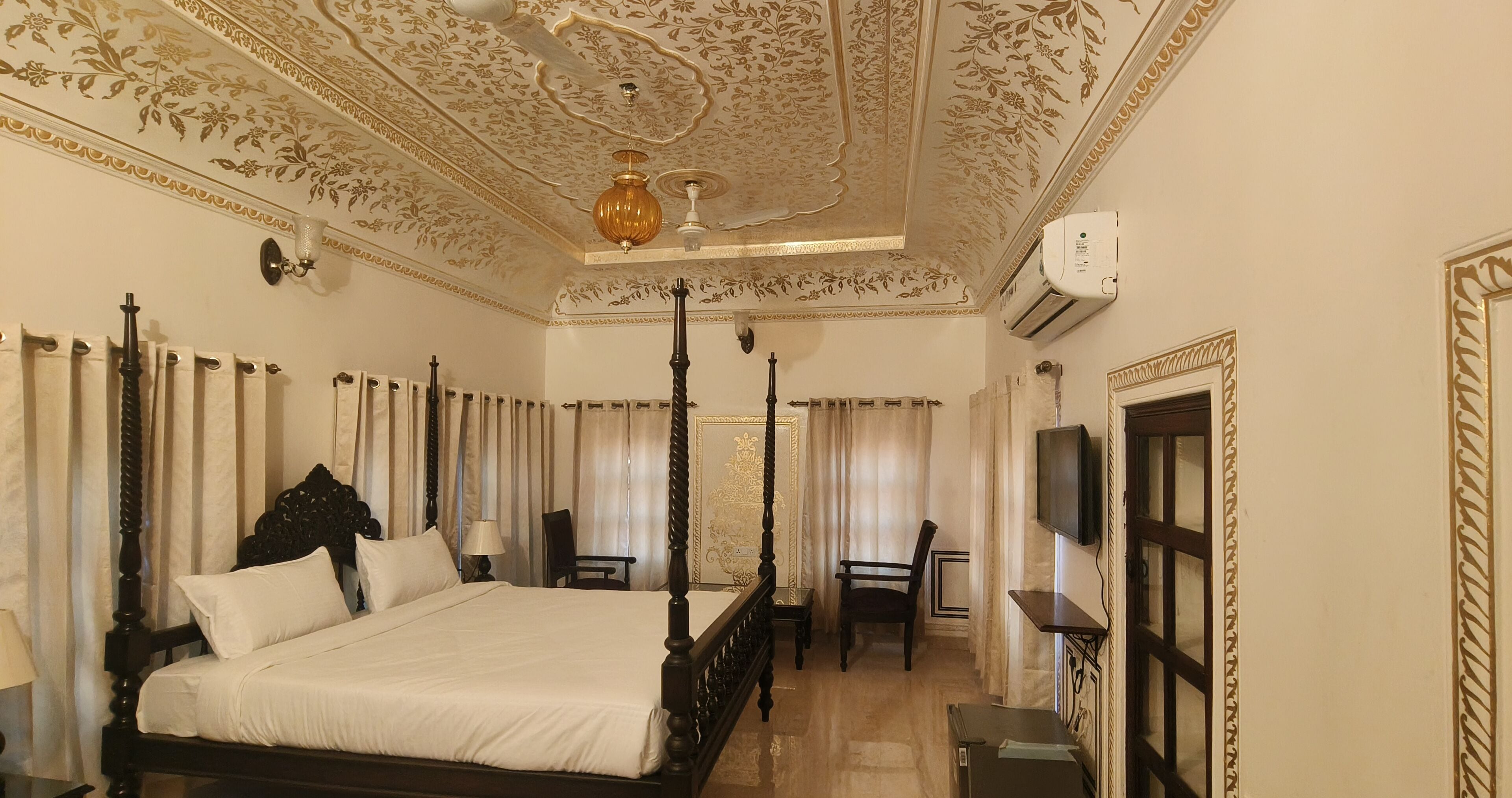 A four-poster bed with a dark wooden frame and white linens, a chandelier, and a television on the wall.