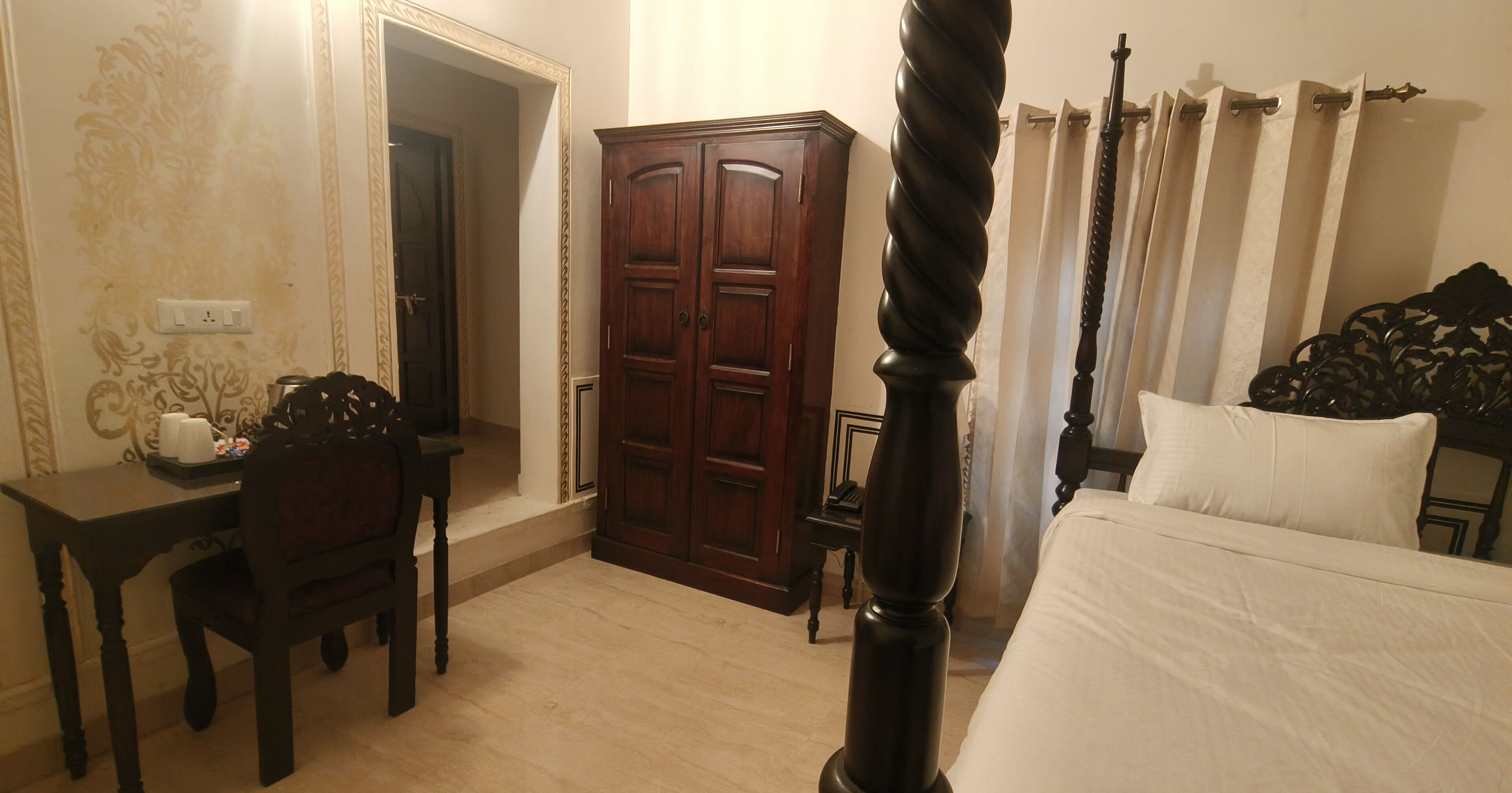 A bedroom with a bed, a wooden wardrobe, a desk with a chair, and a mirror.