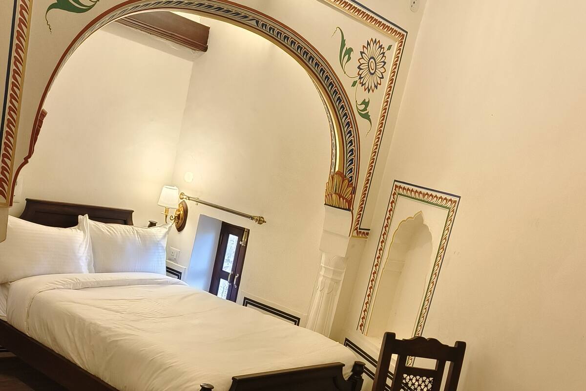 A bedroom with a large bed, a wooden chair, a bedside table with a lamp, and decorative wall patterns.