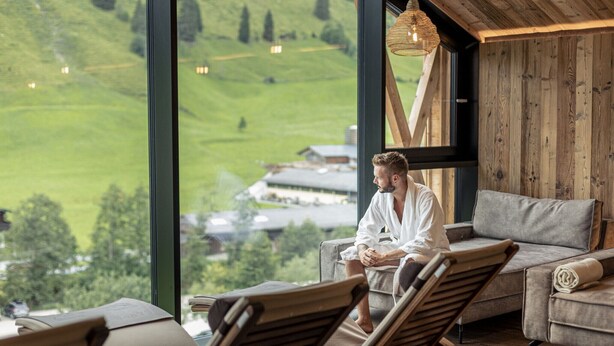 A room with large windows offering a view of a green landscape, a wooden ceiling, and a sofa with a rolled-up blanket.