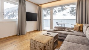 A modern living room with a large sofa, a coffee table, and a flat-screen TV. There are large windows with curtains, offering a view of a snowy landscape. - Spacious apartment with balcony (Rauris)
