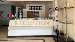 The RIADA HOTEL reception area with a marble counter, clocks displaying time in New York, London, Istanbul, Moscow, and Tokyo, and flags of various countries.