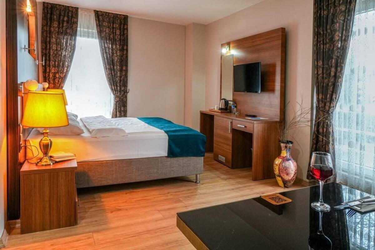 Une chambre d’hôtel comprenant un lit, une table de chevet avec une lampe, un bureau avec une chaise, un téléviseur fixé au mur et une fenêtre avec des rideaux.