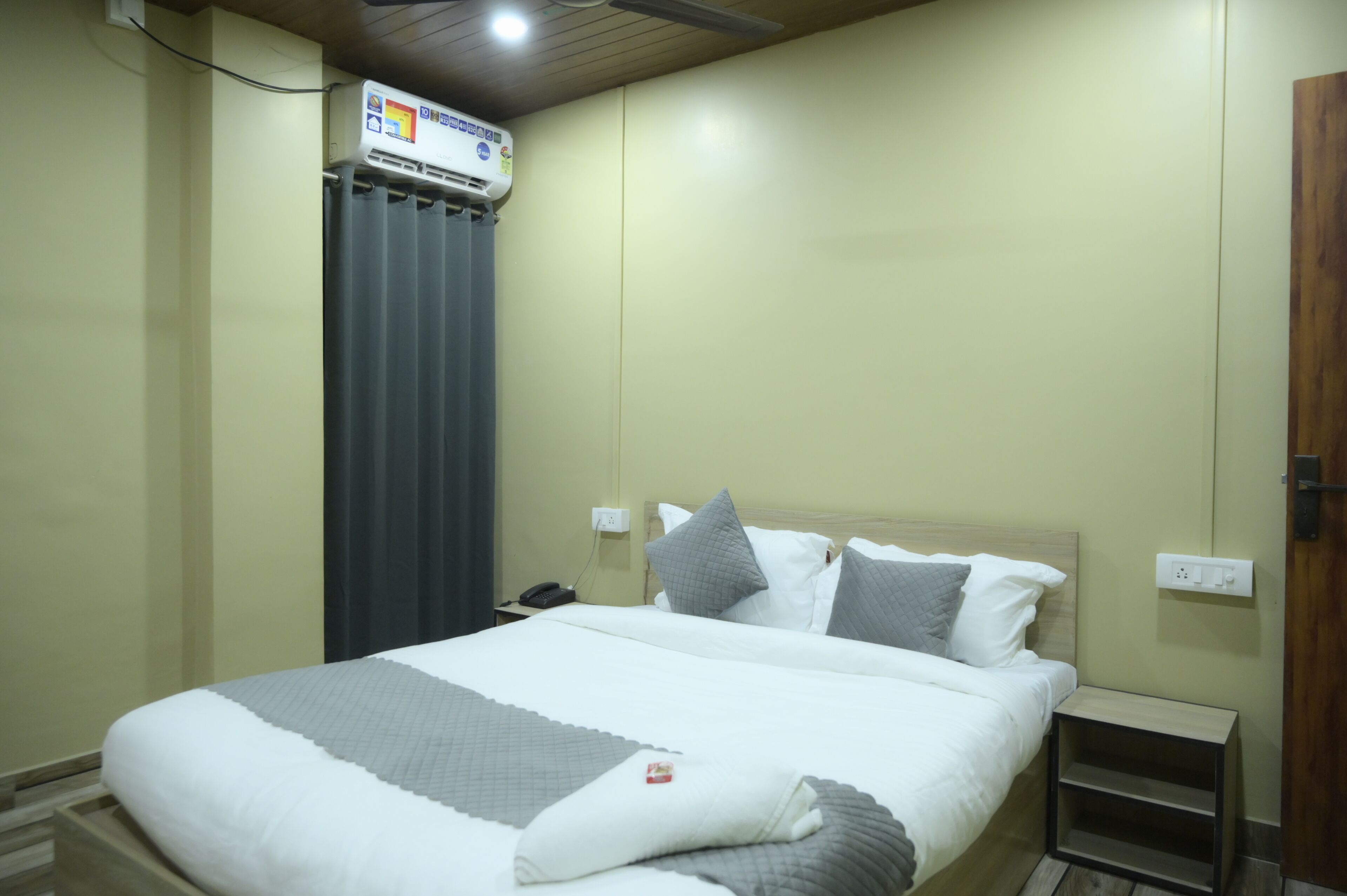 Une chambre d’hôtel avec un lit, une table de chevet, un climatiseur fixé au mur et une porte en bois.