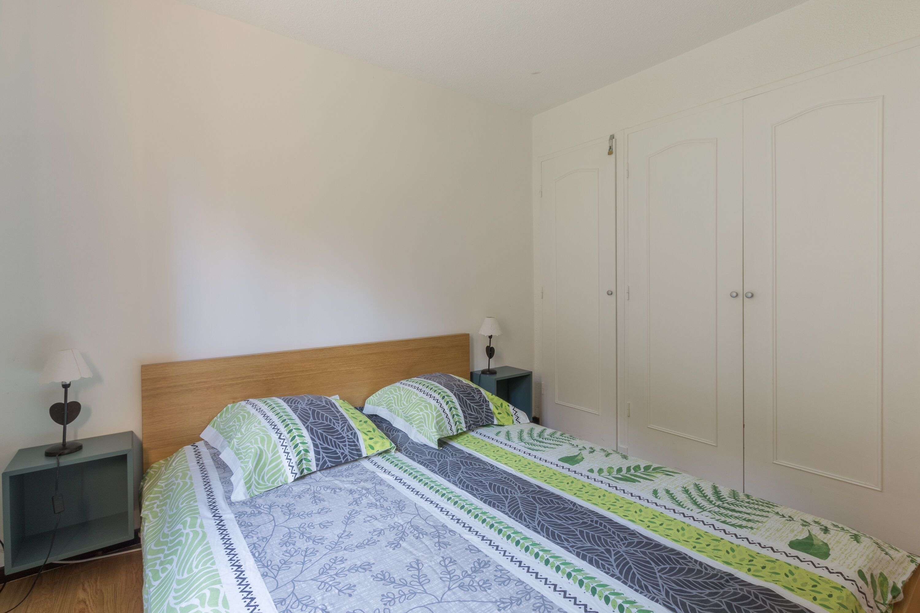 Une chambre à coucher avec une tête de lit en bois, un lit recouvert d’une literie à motifs, deux tables de chevet avec des lampes et une armoire blanche.