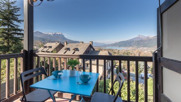 A balcony with a table set for two, overlooking a mountainous landscape with a lake and buildings in the distance.