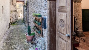 A narrow cobblestone alley with a rustic stone wall, a wooden door with a circular emblem, and a potted plant with purple flowers. - Stunning private villa for 6 people with pool, WIFI, hot tub, A/C, TV, patio and panoramic view (Domaso)