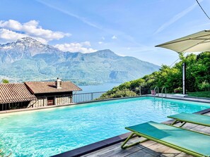 Ein Schwimmbecken mit Blick auf die Berge, eine Liegewiese mit Sonnensegel und ein Gebäude mit einem Ziegeldach.