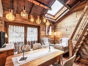 A cozy wooden cabin interior with a dining area, two chairs, a table with a lace tablecloth, a bear-themed wall art, and a wooden staircase.