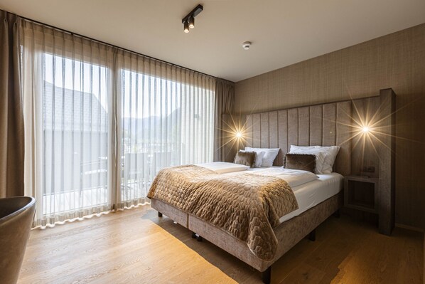Quarto de hotel moderno com uma cama grande, mesa de cabeceira, uma cadeira e vista da cidade através de cortinas translúcidas.