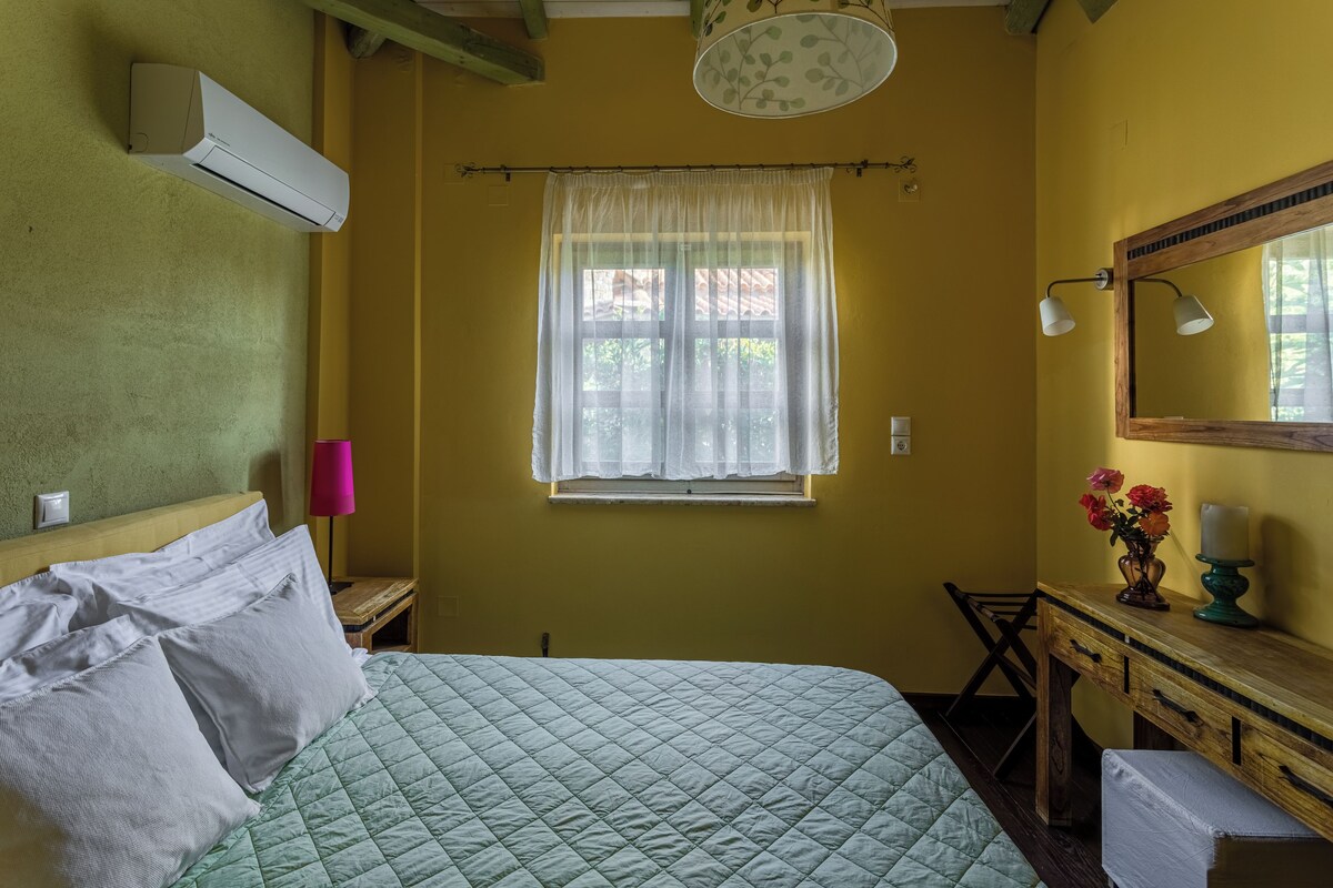 Una habitación con una cama, un ventanal con cortina translúcida, una lámpara de noche, un escritorio de madera, un espejo y un aire acondicionado instalado en la pared.