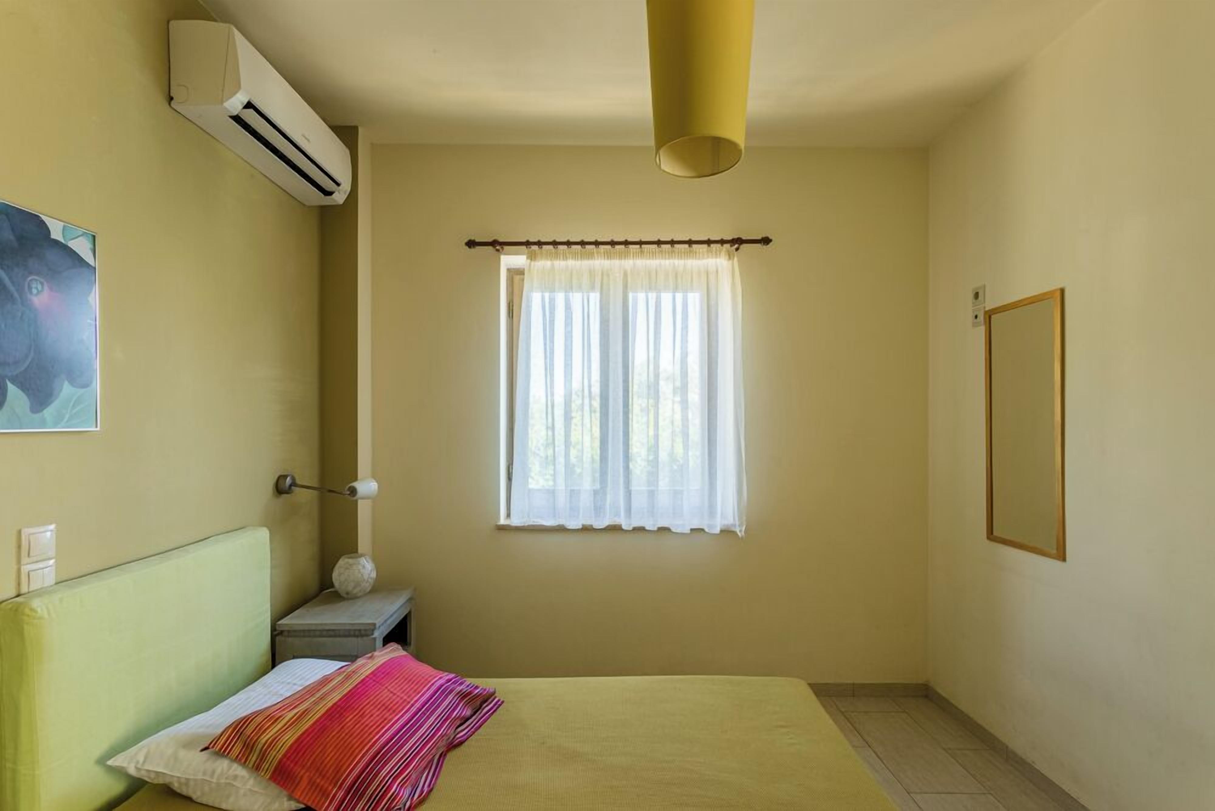 A room with a bed, an air conditioner, a window with curtains, a bedside table, and a wall-mounted picture.
