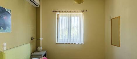 A room with a bed, an air conditioner, a window with curtains, a bedside table, and a wall-mounted picture.