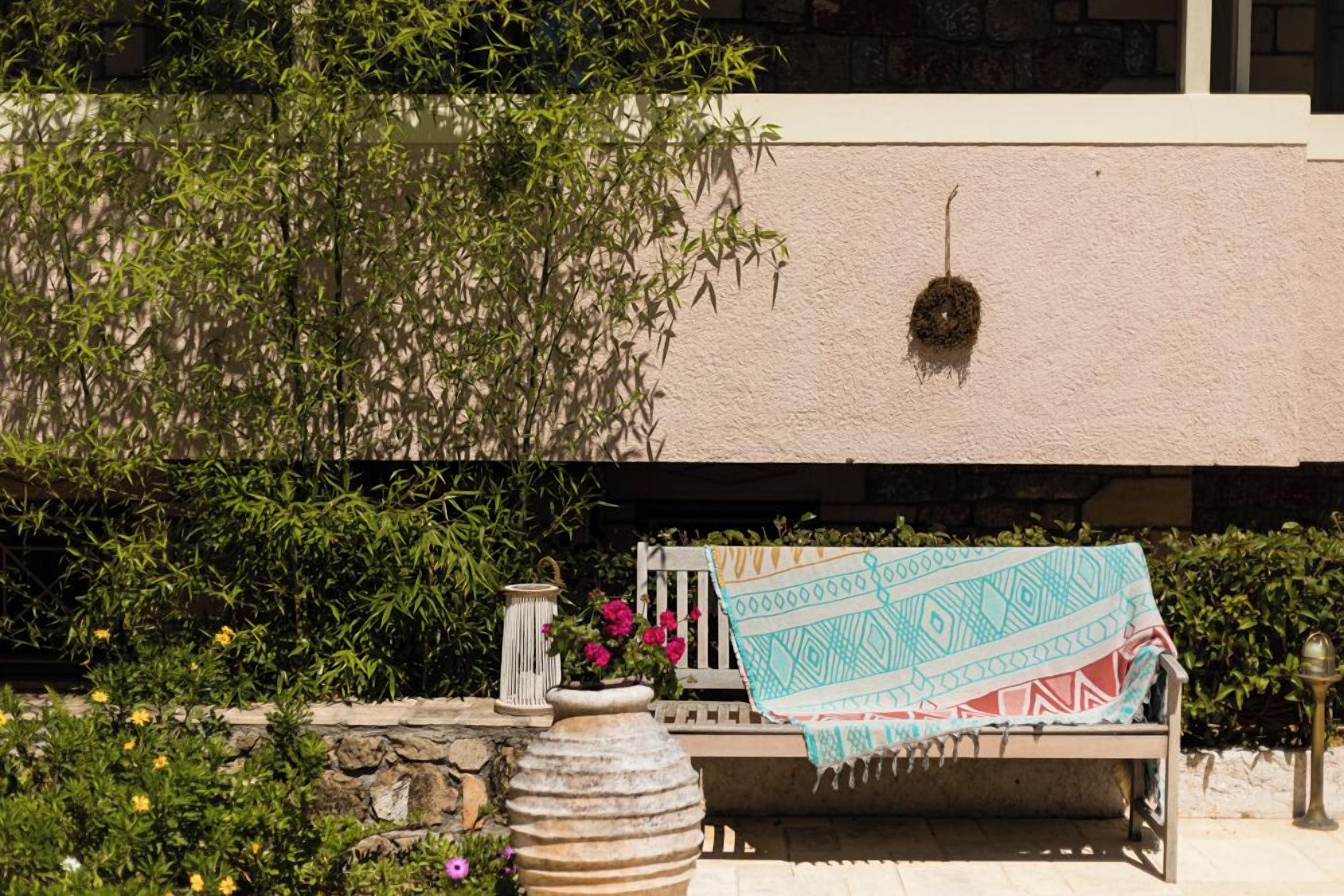 A bench with a patterned throw blanket, a stone vase with flowers, and a lantern.
