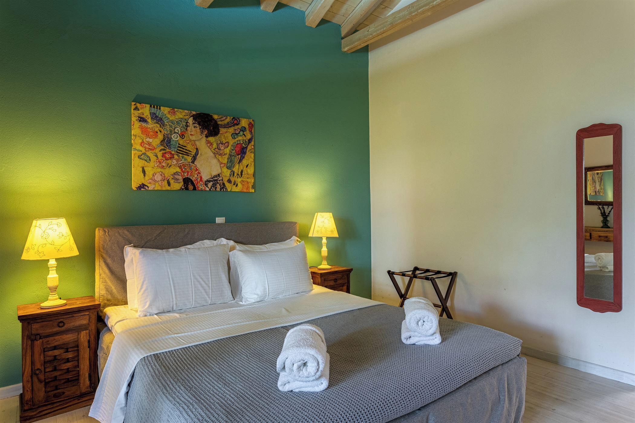 A neatly made bed with white linens, two bedside lamps, a colorful painting above the bed, and a mirror on the wall.