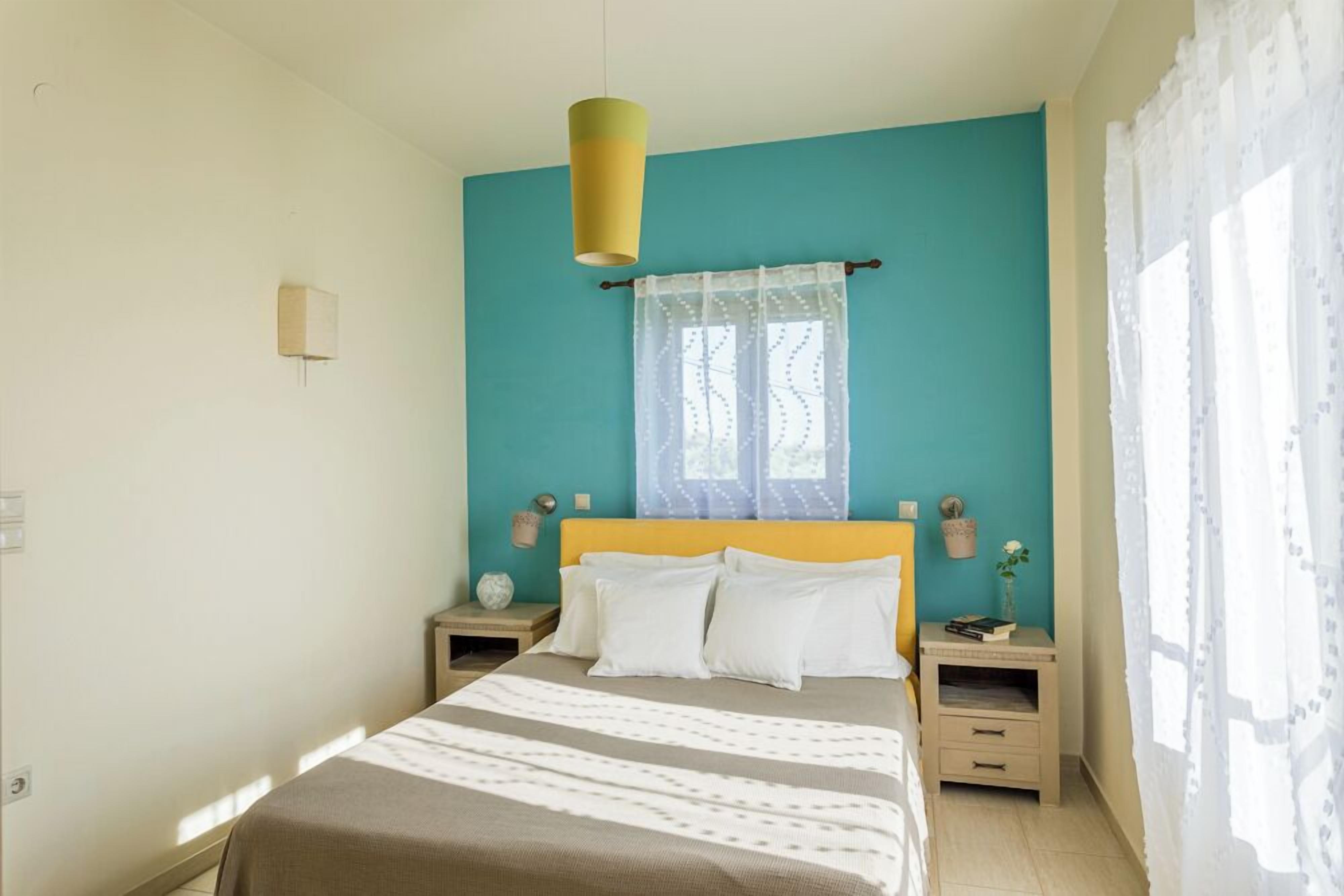 Un dormitorio con una cama, dos mesitas de noche, un ventanal con cortinas translúcidas, una lámpara de pared y una pequeña planta en maceta.