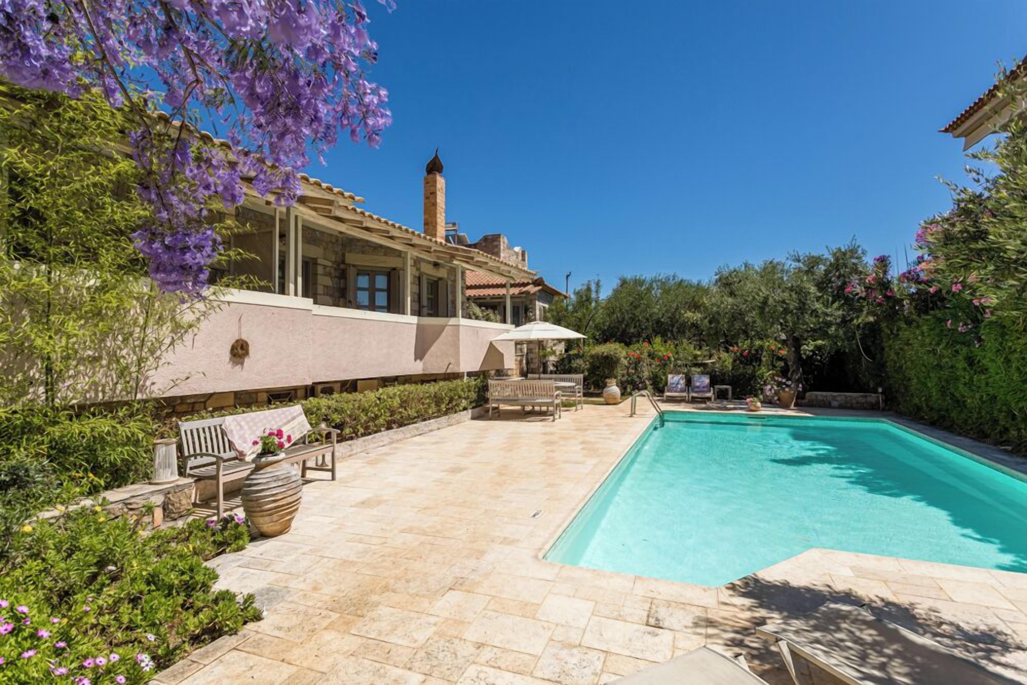 A swimming pool with a patio area, surrounded by greenery and a building with a tiled roof.