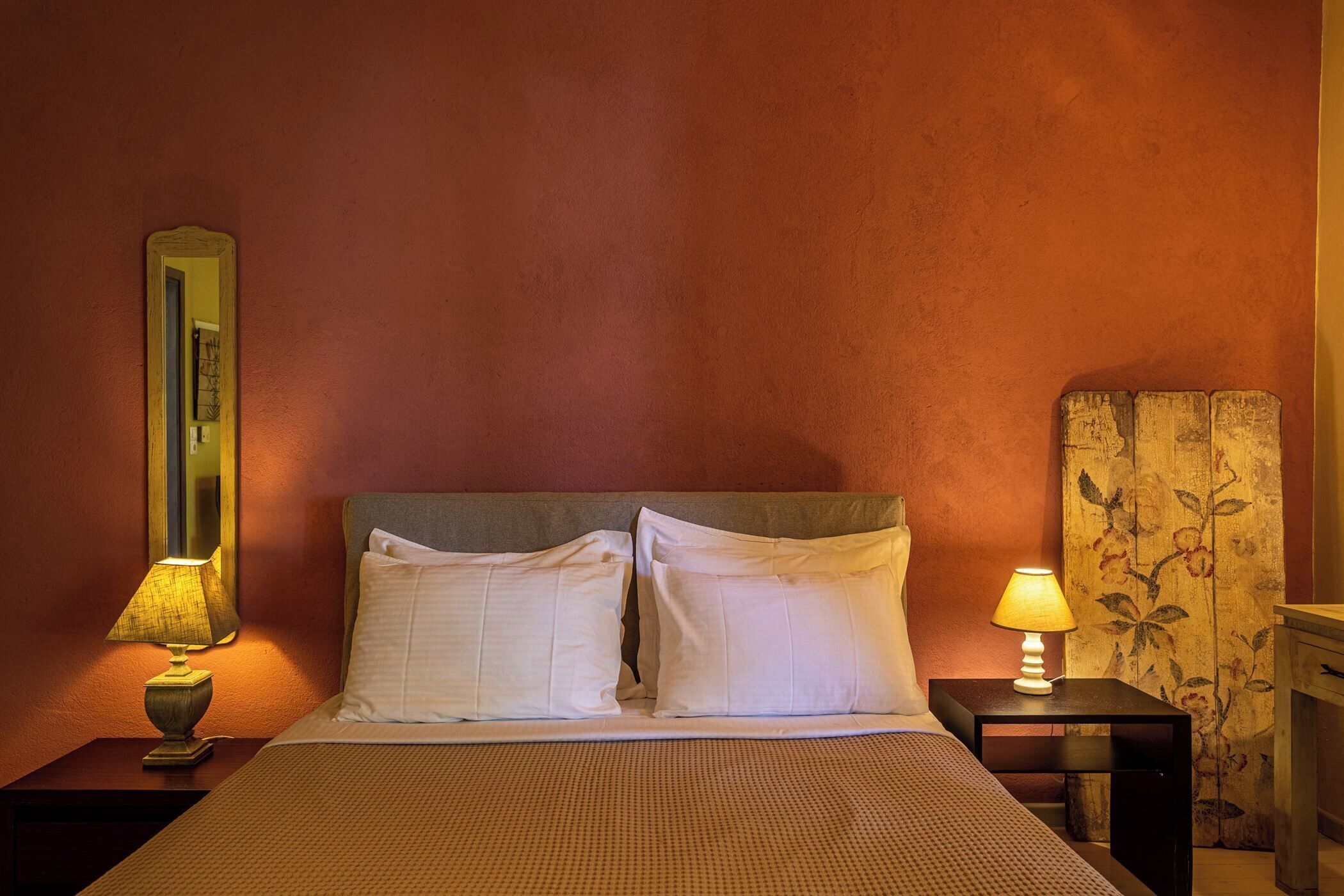 Quarto de hotel com cama, duas luminárias de cabeceira, mesa de apoio, espelho e cortina florida.