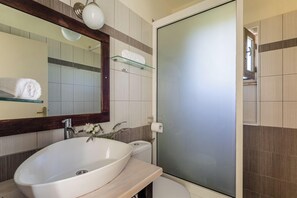 A modern bathroom with a large oval sink, a glass shower enclosure, and a mirror with a wooden frame. - Tria Luxury Residences (West Mani)
