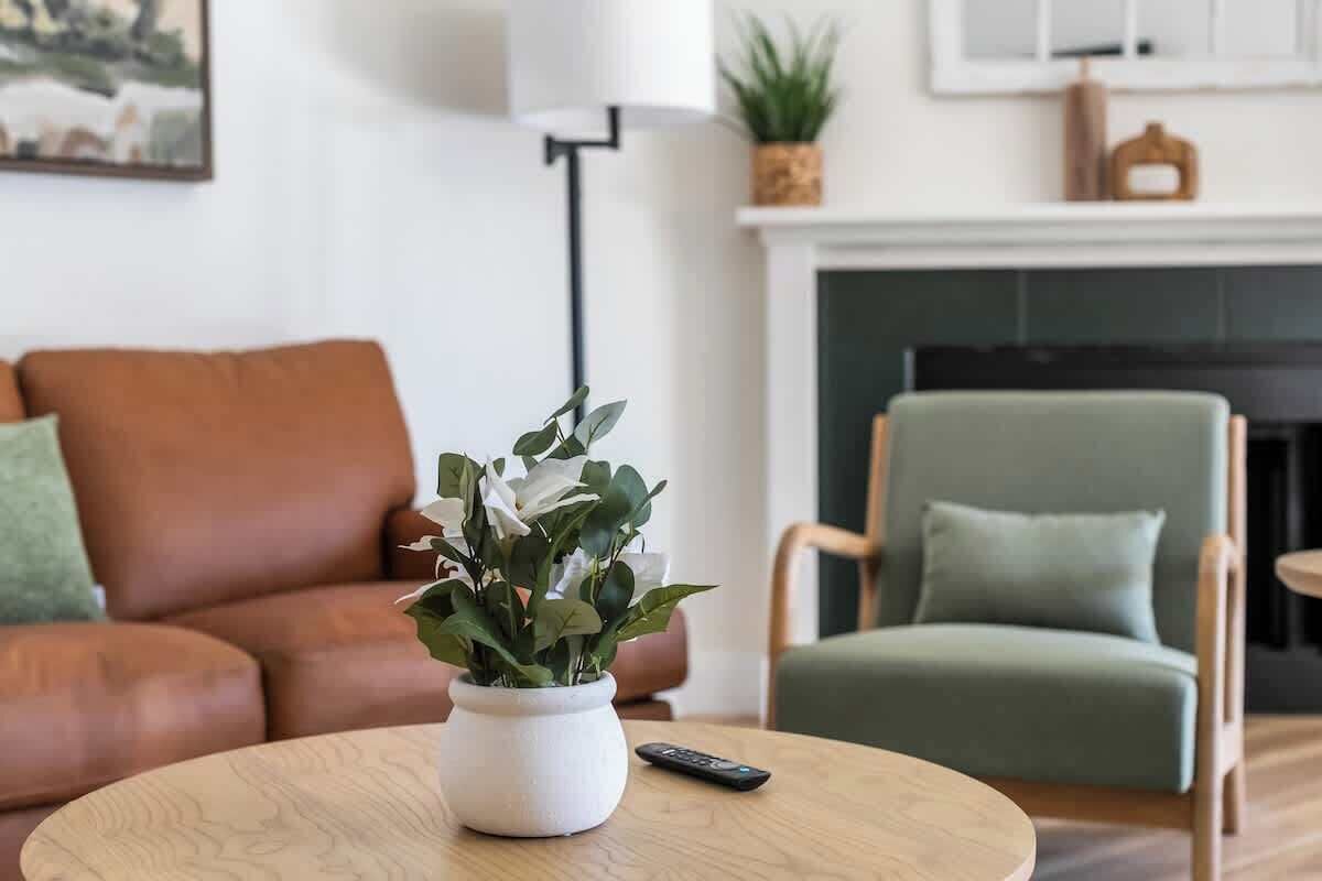 A living room with a brown leather sofa, a green armchair, a round wooden coffee table with a white vase and a remote control, and a fireplace in the background.