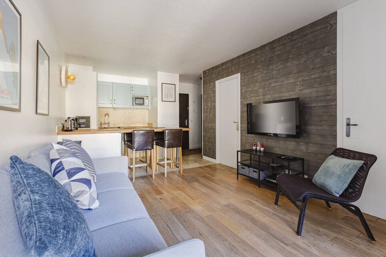 Un soggiorno moderno con un divano grigio, pavimento in legno, una TV montata su una parete scura e un angolo cottura con forno a microonde e sgabelli da bar.