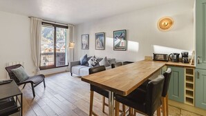 A modern kitchen with a wooden table, a sofa, and a dining area. There are framed pictures on the wall and a window with curtains.