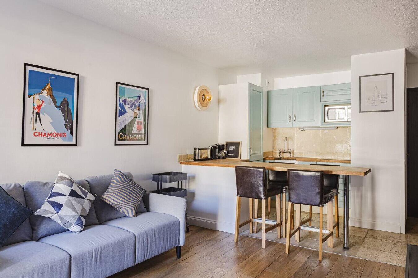 A compact living space with a kitchenette, a sofa with patterned cushions, and two bar stools.