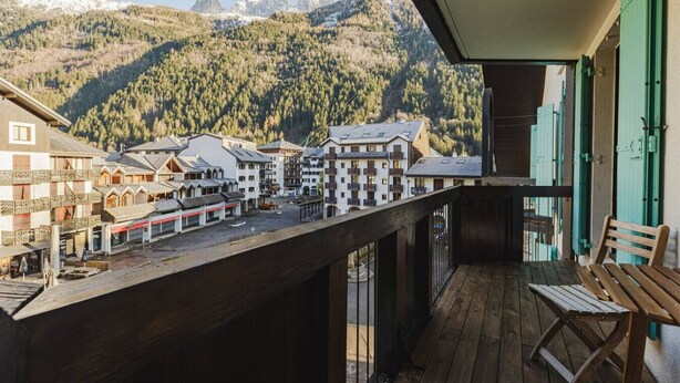 A balcony with a view of a mountain village, wooden furniture, and a building with a sloped roof.