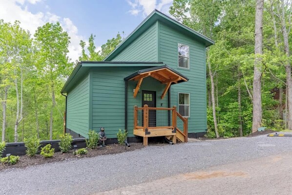 Een groene hut met een houten veranda en een klein terras, omringd door bomen.