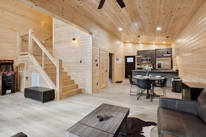 A spacious living area with a wooden staircase, a dining table with chairs, a sofa, and a kitchen area with appliances.